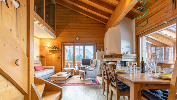 A wooden cabin interior with a dining area, a sofa, a chair, and a staircase.