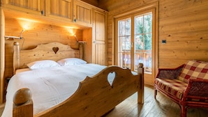 A wooden cabin bedroom with a bed, a wooden headboard with a heart cutout, a wooden bench, and a window with a view of trees.