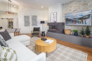 A modern living room with a white sofa, a round wooden coffee table, a fireplace, and a mounted TV displaying a ski resort scene.
