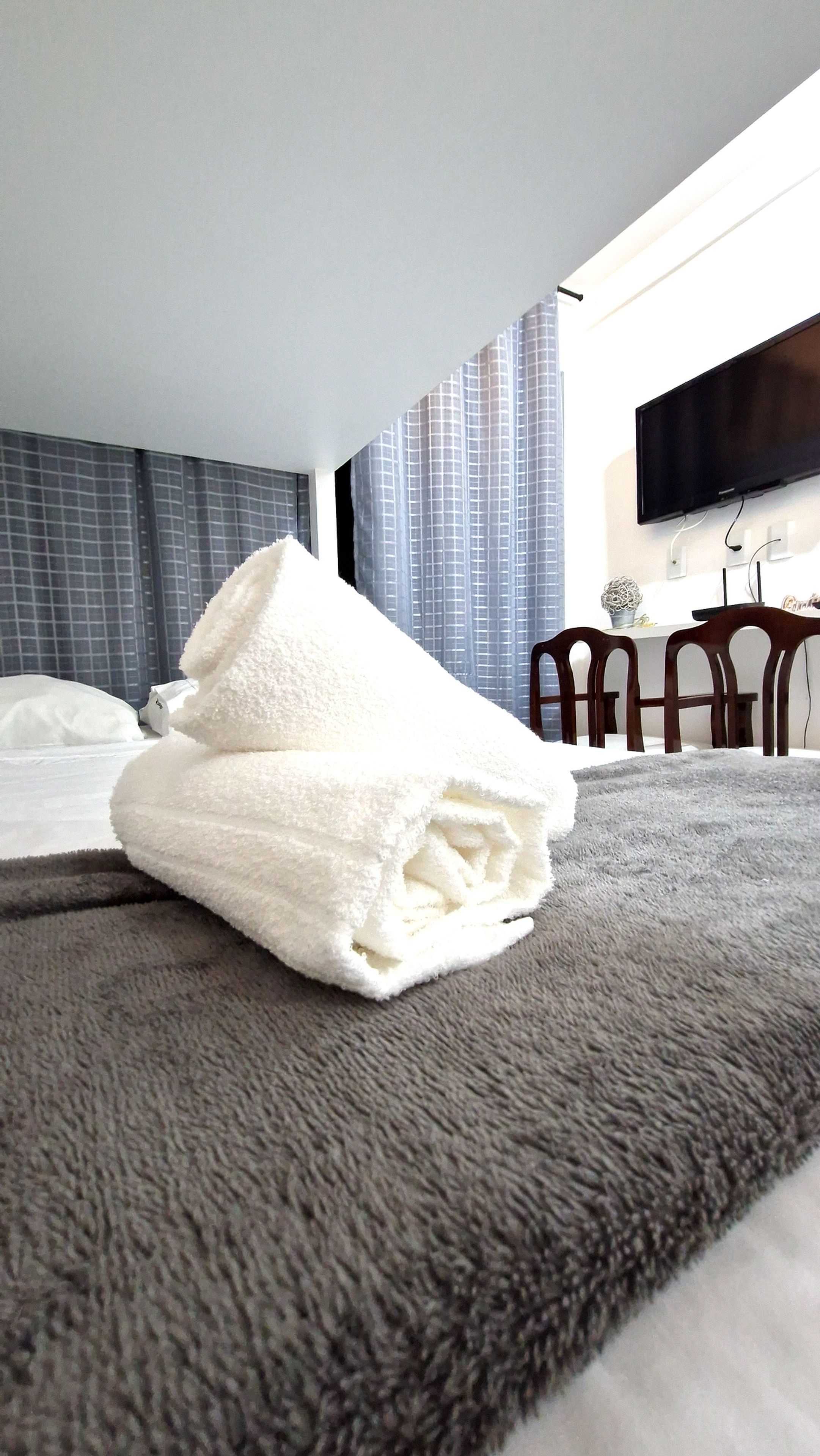 A neatly made bed with a folded white towel, a grey textured rug, and a modern room with a flat-screen TV mounted on the wall.
