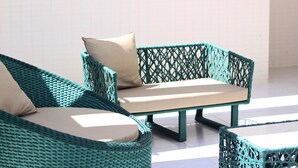 A wicker sofa with a cushion and a matching armchair, both with a beige cushion, in a sunlit room with a tiled floor.