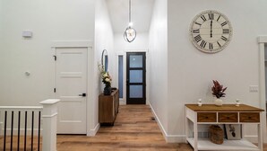 A hallway with a wooden floor, a white door, a mirror, a vase with flowers, a clock, and a small table with decorative items.
