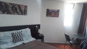 A hotel room with a bed, a desk, a chair, and two paintings on the walls. - Daya apartamentos  (Santiago)
