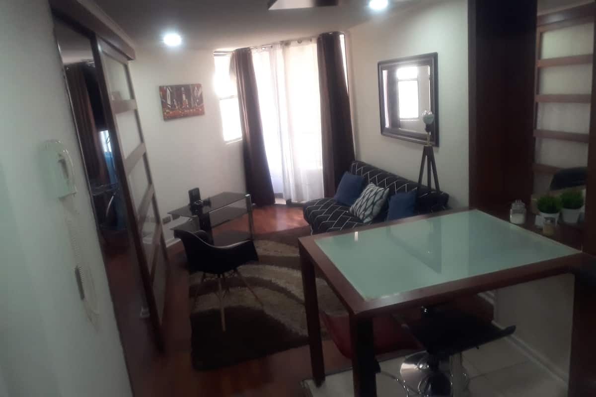 Un espacio habitable compacto con una mesa de comedor de vidrio, un sofá con cojines estampados, una lámpara de pie y un cuadro en la pared.
