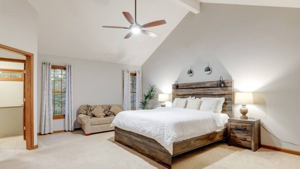Un dormitorio con cama de madera, ventilador de techo, mesita de noche con lámpara, ventana con cortinas y sofá.