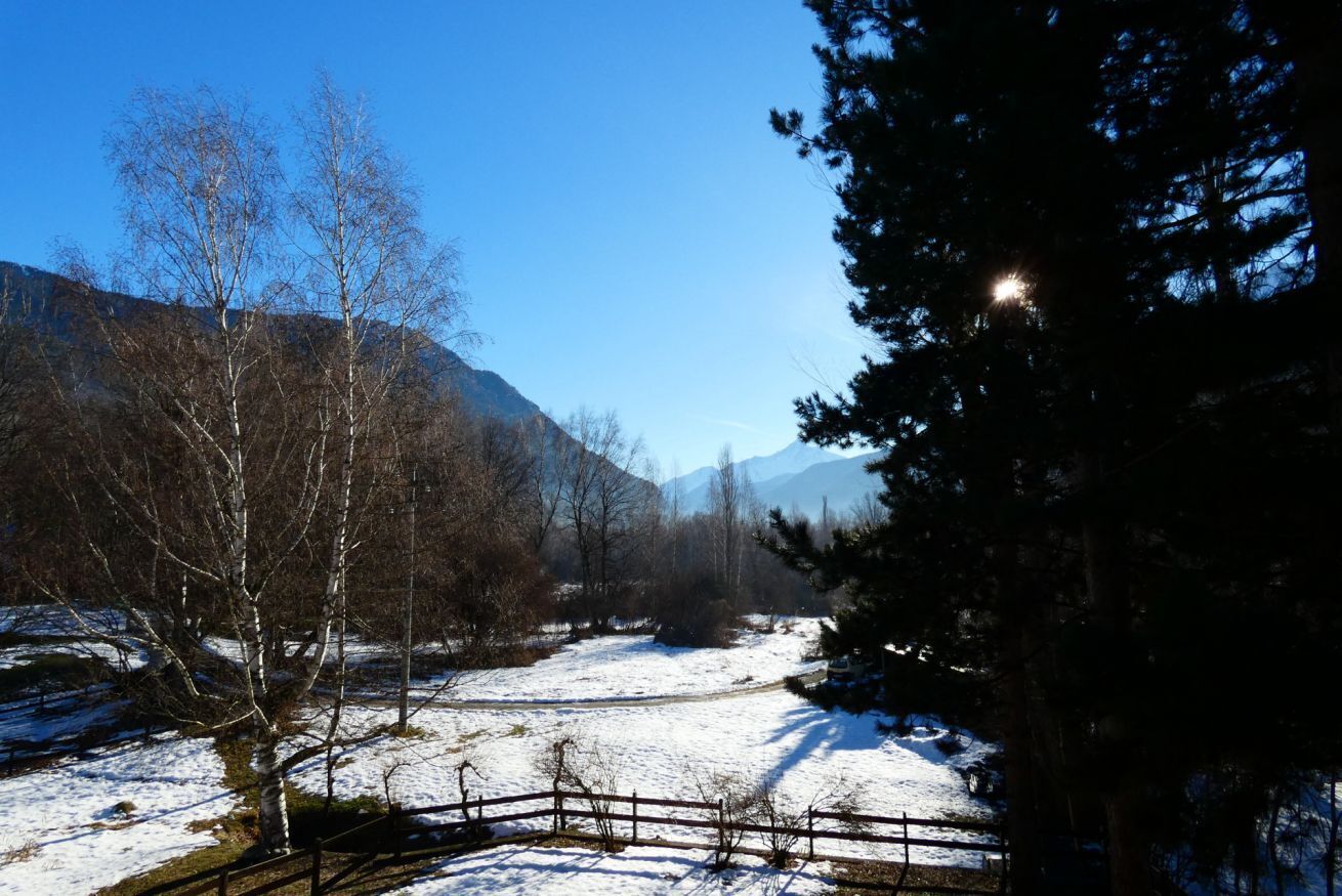 Schneebedeckte Landschaft mit Holzzaun, Bäumen und Bergen im Hintergrund.