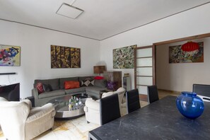 A living room with a sofa, coffee table, and chairs, a dining table with a vase, and artwork on the walls.
