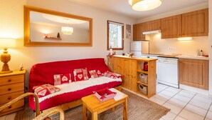 A cozy living room with a red sofa, wooden furniture, a kitchen with wooden cabinets, and a large mirror. - Le Refuge du Chevreuil - Pistes accessibles à pieds (Les Angles)