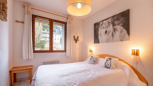 A bedroom with a large bed, a wooden bench, a window with curtains, a wall-mounted deer head, and a wolf painting. - Le Refuge du Chevreuil - Pistes accessibles à pieds (Les Angles)