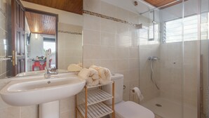 Un baño con lavabo, inodoro y ducha. Hay un perchero con toallas y un estante.