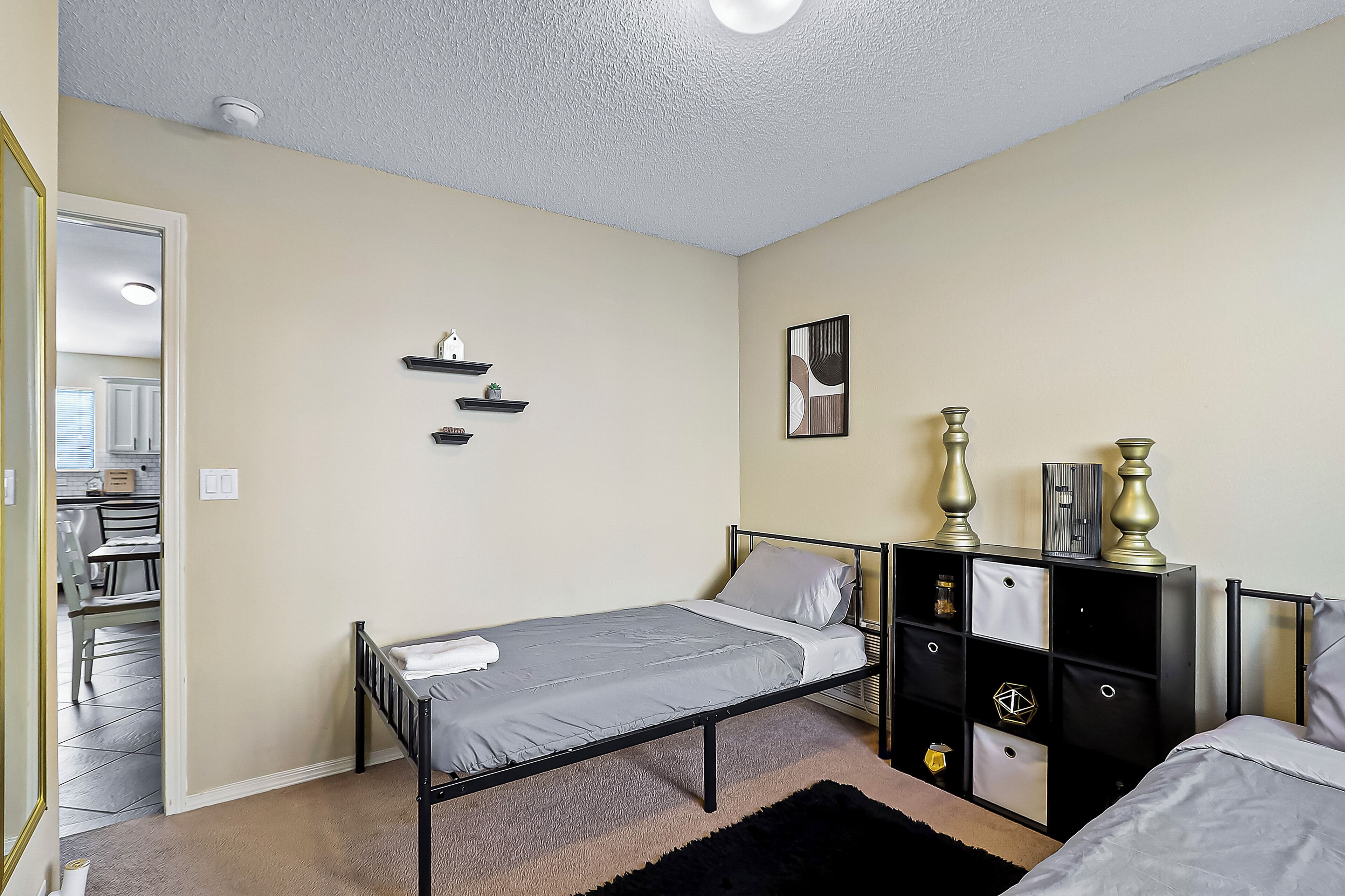 A bedroom with a single bed, a black metal dresser with drawers, a small shelf unit, and a framed picture on the wall.