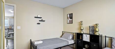 A bedroom with a single bed, a black metal dresser with drawers, a small shelf unit, and a framed picture on the wall.