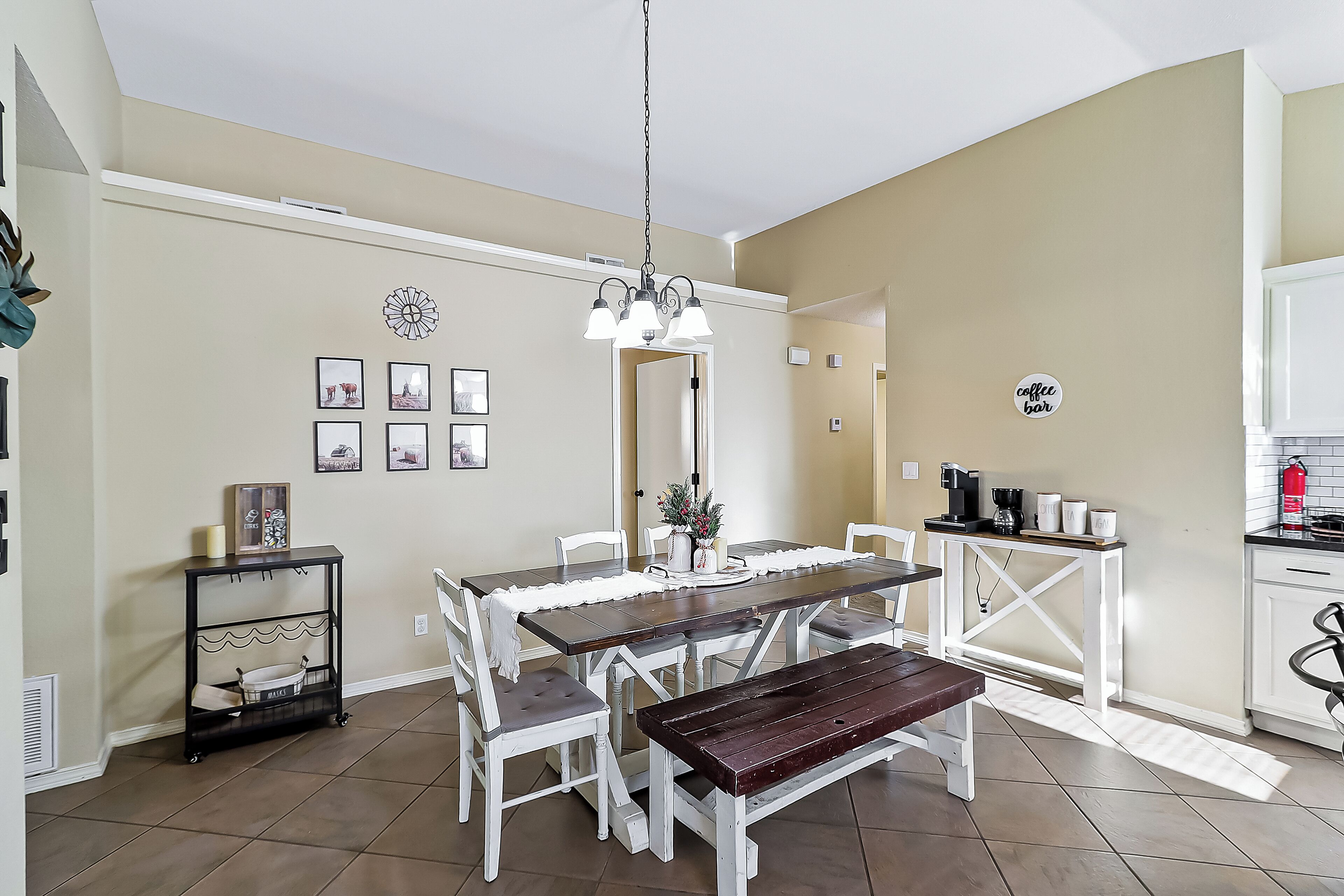 Un comedor con mesa de madera, sillas blancas, un banco, una lámpara colgante y una zona de cocina con estación de café.