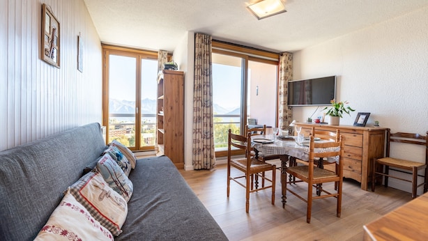 Una sala de estar acogedora con un sofá, una mesa de comedor con sillas, un televisor y vistas a las montañas a través de ventanas amplias.