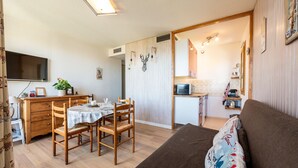 Una sala de estar acogedora con un sofá marrón, una mesa de comedor con sillas, un televisor montado en la pared y una cocina visible a través de un vano abierto.