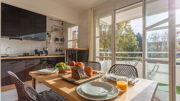 A modern kitchen with dark cabinets, a wooden table set for breakfast, and a large window offering a view of trees and buildings. - Place Cosy - Charmant appt pour 4 (Courcouronnes)