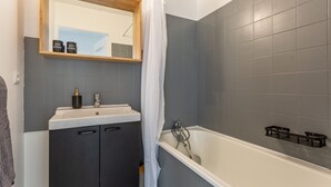 A bathroom with a white bathtub, a dark vanity, and a mirror above the sink. - Place Cosy - Charmant appt pour 4 (Courcouronnes)