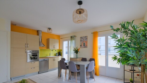 A modern kitchen with a dining area, featuring a central island, built-in appliances, and a large window with curtains. - La Balnéaire - Vue mer, à 50 m de la plage et de la baie (Saint-Nazaire)