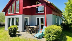 Una casa de dos pisos con fachada roja y detalles blancos, que cuenta con un patio con mobiliario exterior y un pequeño jardín.