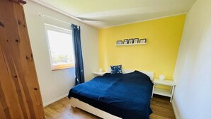Un dormitorio con una cama, una ventana con cortinas, una estantería con fotografías y suelo de madera.