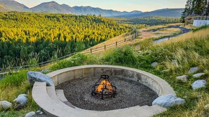 A circular fire pit with a metal grate, surrounded by a concrete border, set in a grassy area with a view of a forested valley and mountains in the background. - Charming Suncadia Condo with Private Balcony, Mountain Views and Pool Access (Cle Elum)