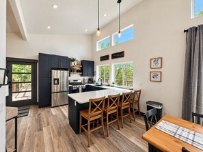 Une cuisine moderne dotée d’un îlot central, de meubles de rangement aux couleurs sombres, d’appareils électroménagers en acier inoxydable et d’un coin repas avec des chaises en bois.