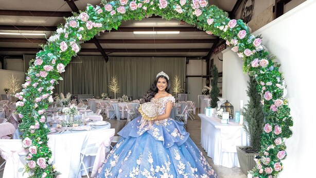 A person in a blue gown with floral patterns standing under a floral archway in a room with tables set for an event. - Countryside Texas Sanctuary Enjoy Sunrises & Sunsets, Grill, Firepit & Longhorns (Pattison)