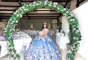 A person in a blue gown with floral patterns standing under a floral archway in a room with tables set for an event.