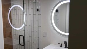 A bathroom with a white tiled wall, a glass shower enclosure, a white sink with a black faucet, and two round mirrors with illuminated frames.