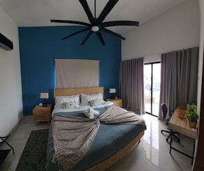 Ein Schlafzimmer mit einem Bett, einem Deckenventilator, einem Nachttisch mit Lampe, einem Schreibtisch mit Stuhl und einem Fenster mit Vorhängen.