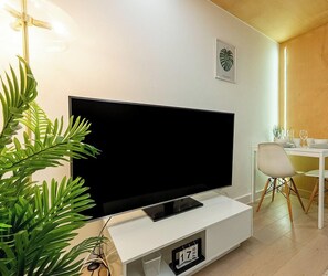 A modern living room with a flat-screen TV on a white stand, a dining table with chairs, and a potted plant.
