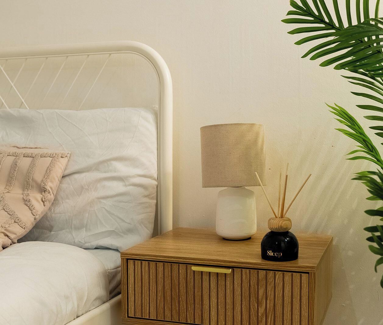 Una cama con estructura metálica blanca, almohada color beige, mesita de noche con lámpara y difusor de aroma, y una planta verde.