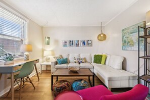 A modern living room with a white sofa, a wooden coffee table, and a desk with a chair. There are decorative pillows and a hanging lamp.