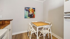 Una cocina pequeña con una mesa de madera y sillas blancas, un colorido cuadro de vaca en la pared y un refrigerador blanco.