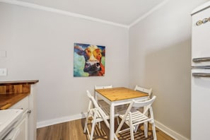 A small kitchen with a wooden table and white chairs, a colorful cow painting on the wall, and a white refrigerator.