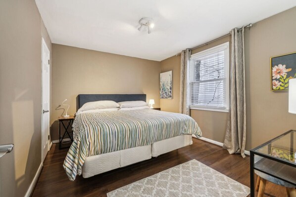 A bedroom with a bed, bedside table, lamp, window with curtains, and a painting on the wall.