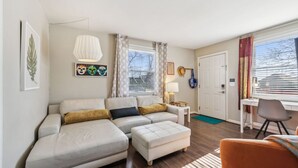 Una sala de estar moderna con un sofá, un reposapiés y una pequeña mesa con una lámpara. Se ve una puerta y una ventana con cortinas.