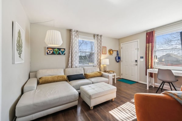 A modern living room with a sofa, ottoman, and a small table with a lamp. There is a door and a window with curtains.