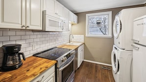 Cocina compacta con armarios blancos, una placa de inducción negra, microondas empotrado, horno de acero inoxidable, lavaplatos y lavadora blanca.