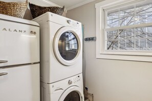 Een kleine wasruimte met een voorlader wasmachine en droger, een raam met uitzicht op kale bomen, en een witte deur.