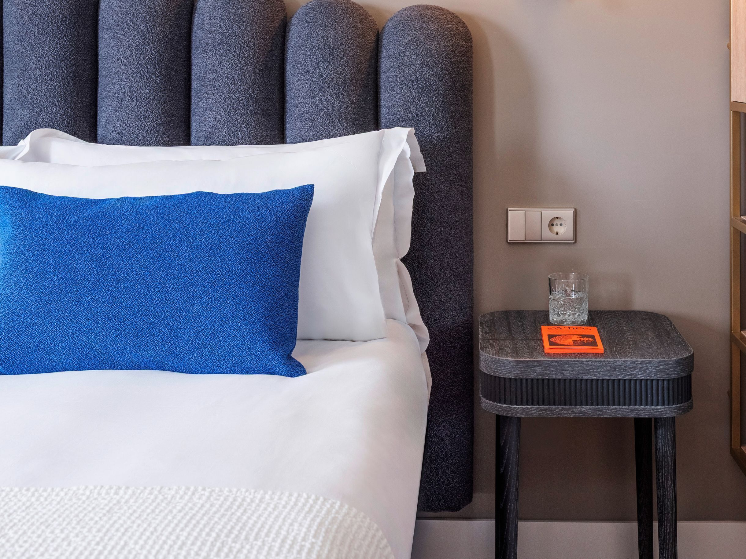 Ein ordentlich bezogenes Bett mit weißen und blauen Kissen, ein kleiner Nachttisch mit einem Glas Wasser und einer Karte, sowie ein an der Wand befestigter Lichtschalter.