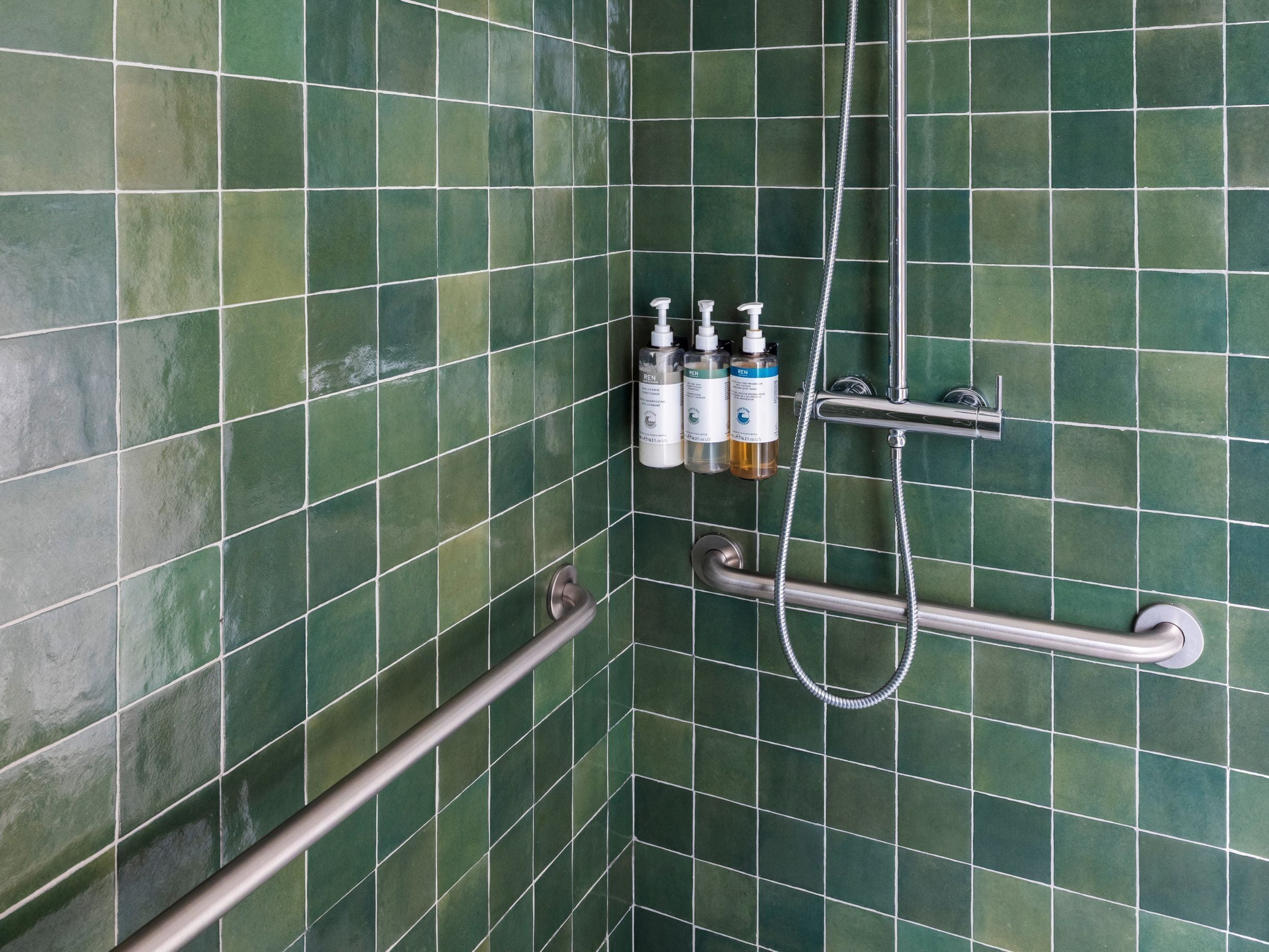 Un espace douche avec des carreaux verts, une barre d’appui et trois bouteilles de shampoing et d’après-shampoing.