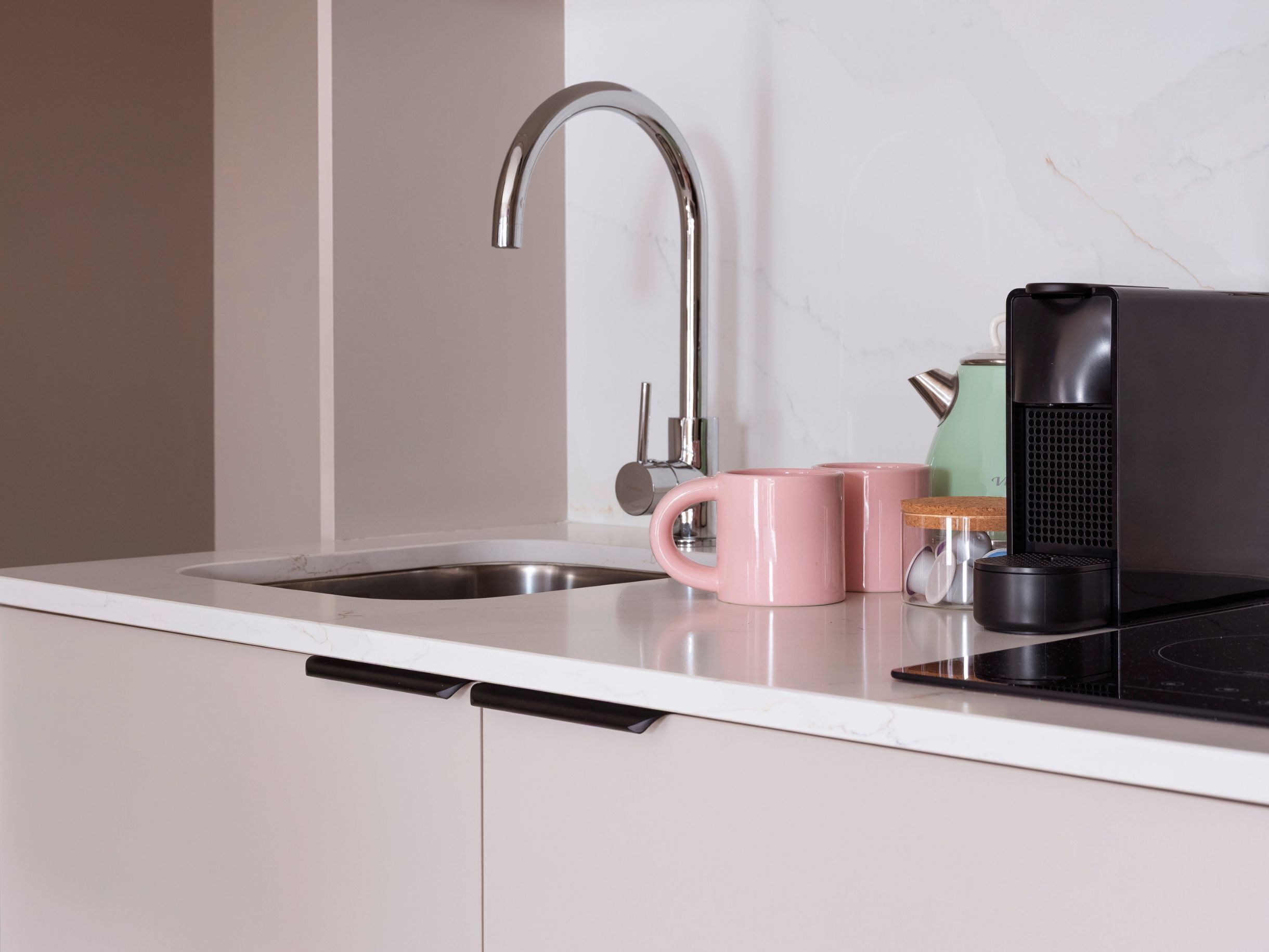 Une cuisine avec un évier en acier inoxydable, une bouilloire électrique noire et une tasse rose posée sur le comptoir.