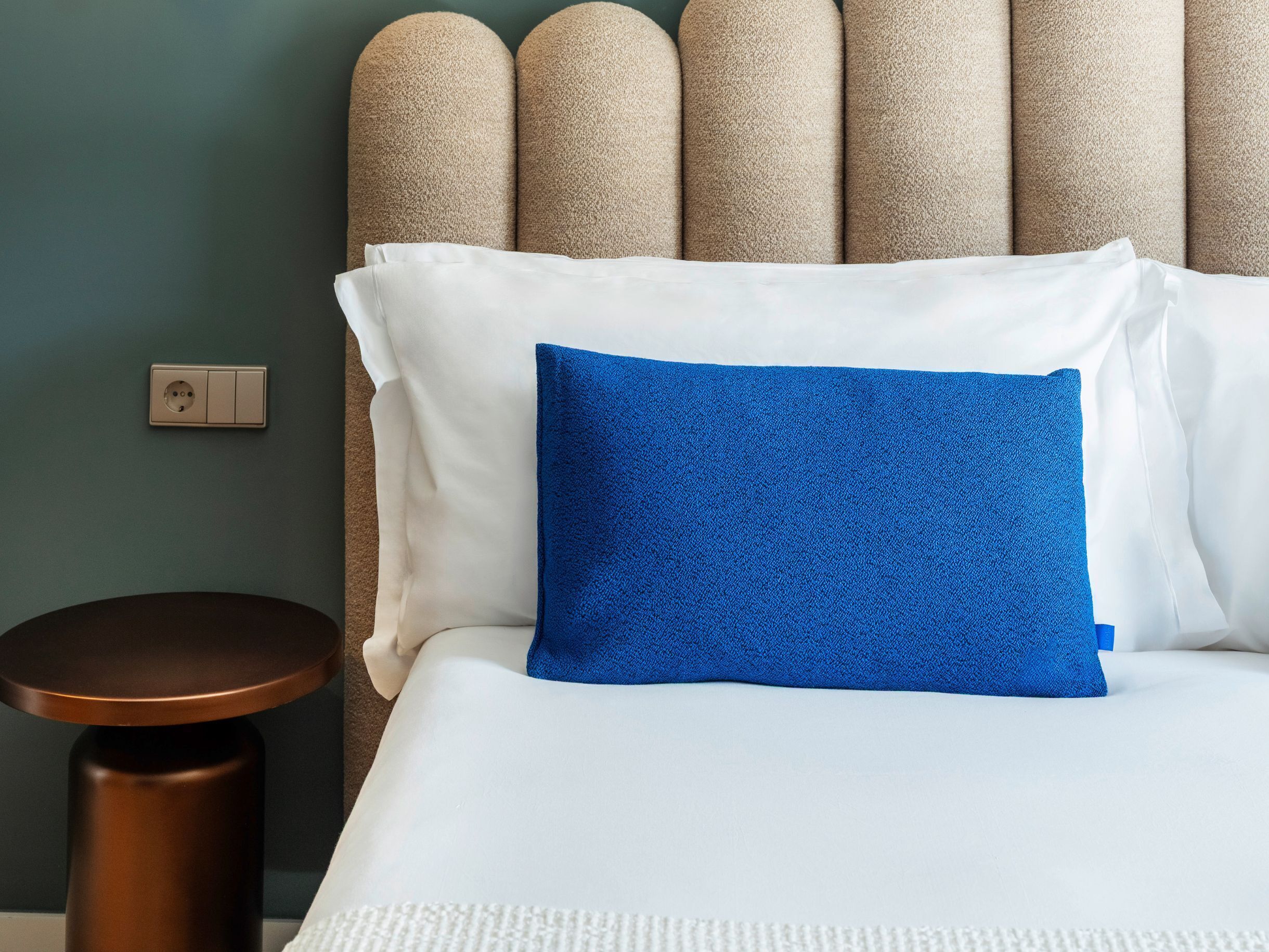 A neatly made bed with white and blue pillows, a round wooden side table, and a textured headboard against a teal wall.