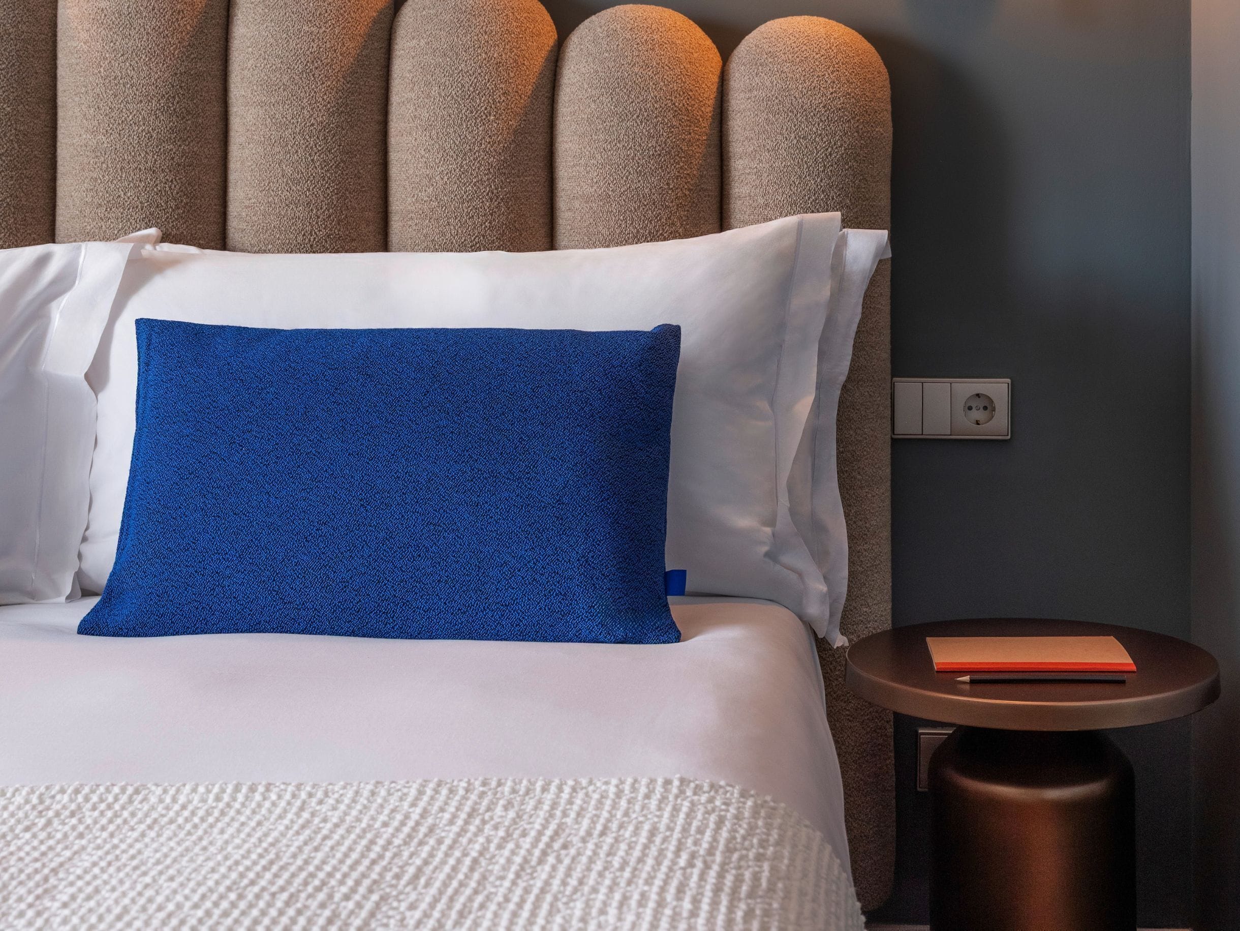 Un lit bien fait, avec des oreillers blancs et bleus, une couverture blanche à texture et une tête de lit marron. Une petite table ronde, sur laquelle se trouve un livre et une prise électrique, est visible sur la droite.