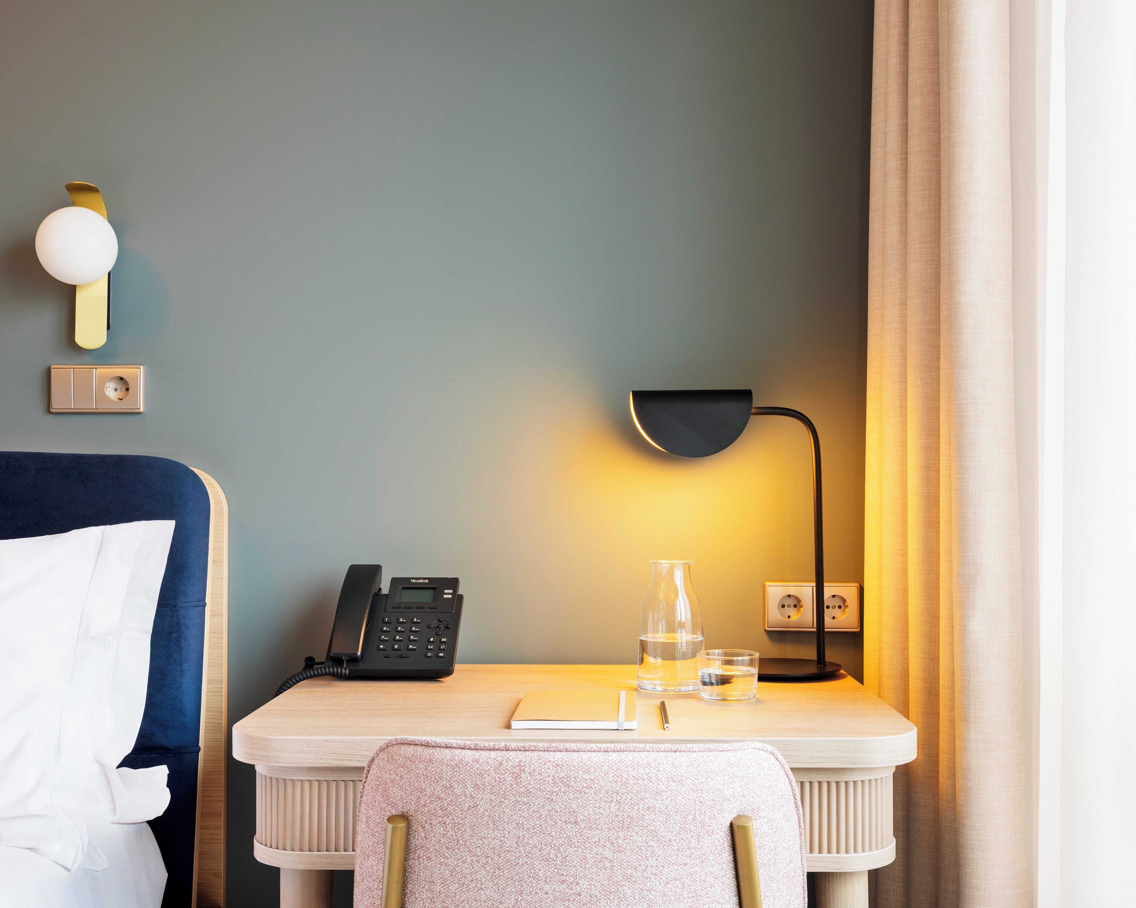 Et hotelværelse med en seng, et skrivebord med telefon, lampe, et glas vand og en notesbog.