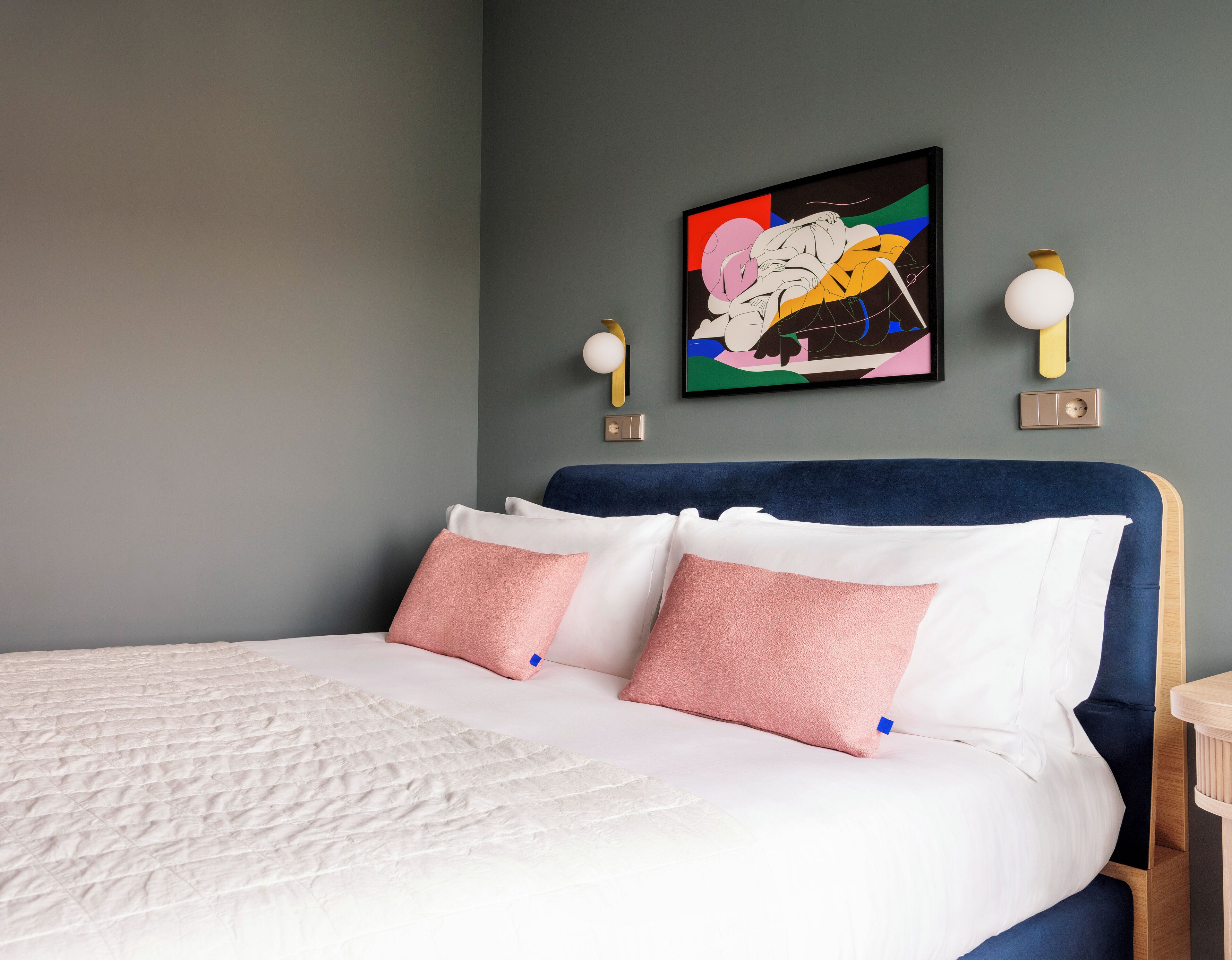Una cama bien hecha con almohadas blancas y rosadas, un cabecero azul y un cuadro abstracto lleno de color encima.