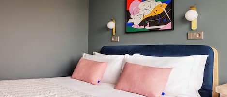Una cama bien hecha con almohadas blancas y rosadas, un cabecero azul y un cuadro abstracto lleno de color encima.