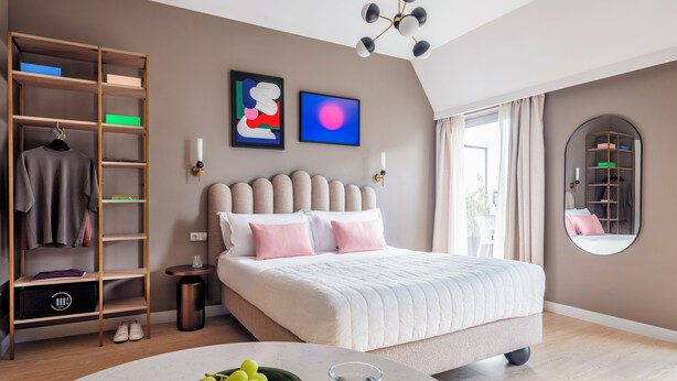 Une chambre moderne avec un grand lit, une armoire intégrée, une table ronde sur laquelle se trouve un saladier rempli de fruits et un verre, ainsi qu’une grande fenêtre avec des rideaux.