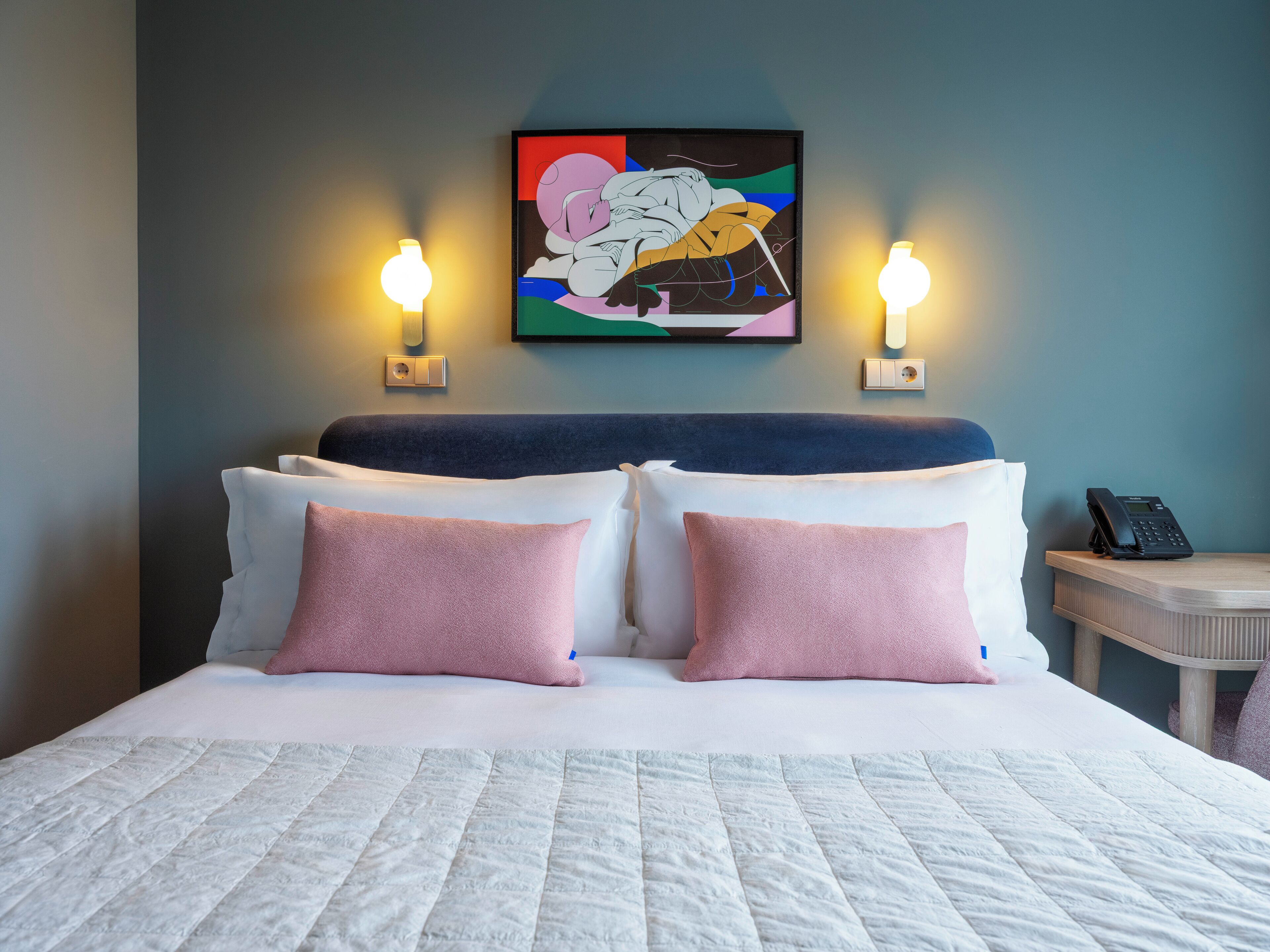 Une chambre d’hôtel avec un lit, deux lampes de chevet, une table de chevet avec un téléphone et un tableau abstrait aux couleurs vives accroché au mur.